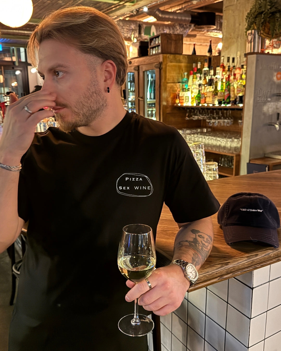 Man in a bar wearing a Pizza & Sex & Wine Organic T-shirt, holding a glass of wine.