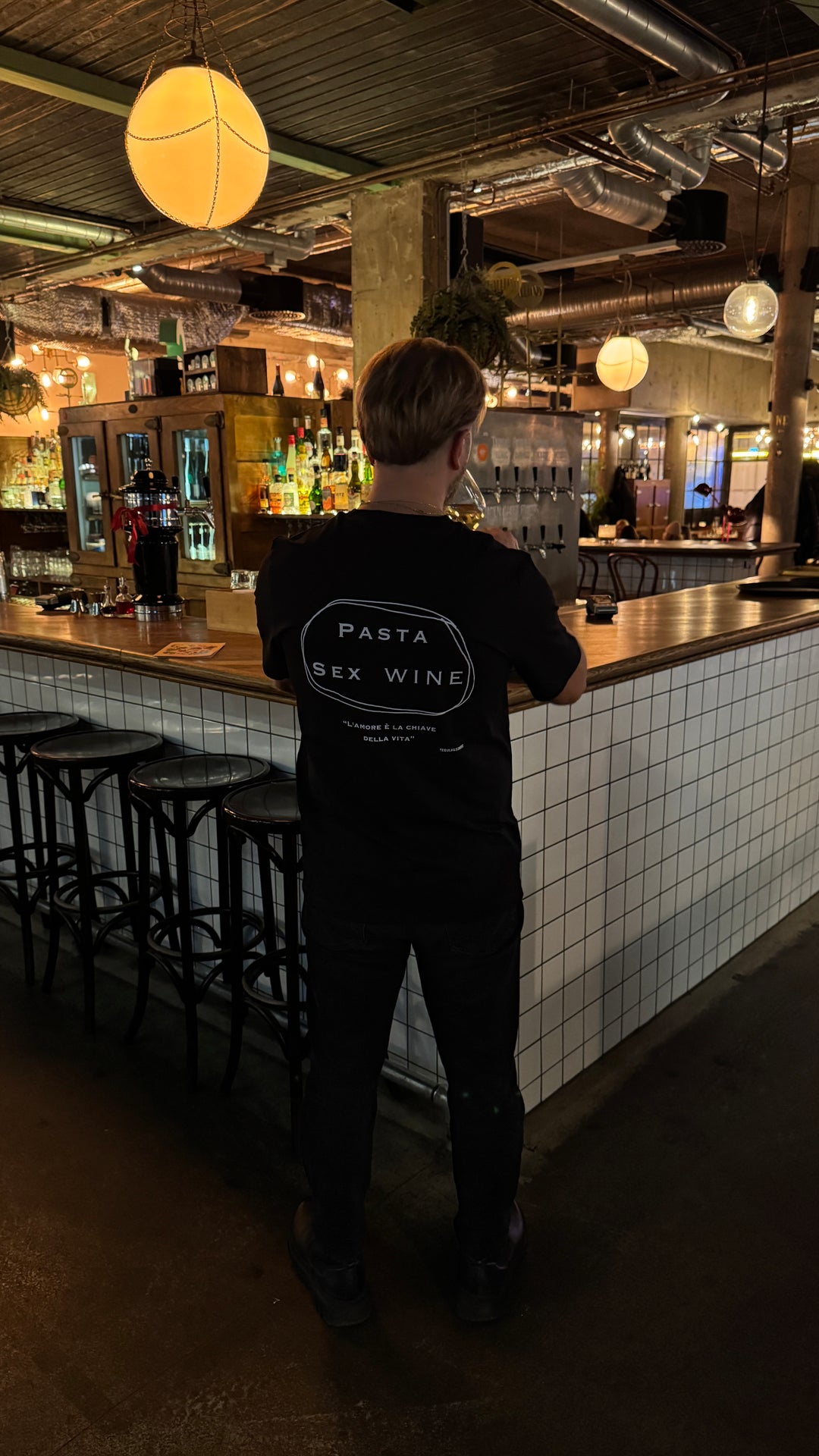 Person wearing a Pasta & Sex & Wine Organic T-shirt in a bar setting, showcasing the trendy design and relaxed vibe.