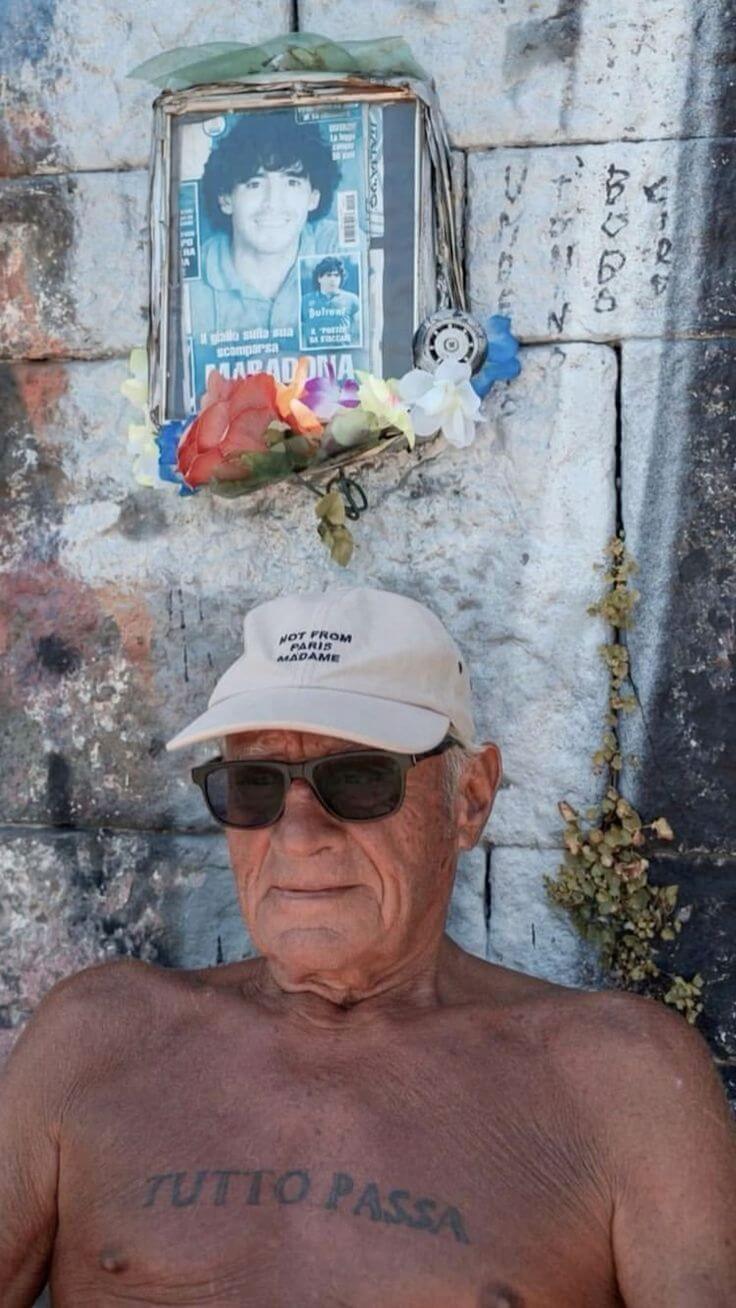 Man wearing sunglasses and a cap with a tattoo on his chest, standing in front of a wall with a portrait and flowers.