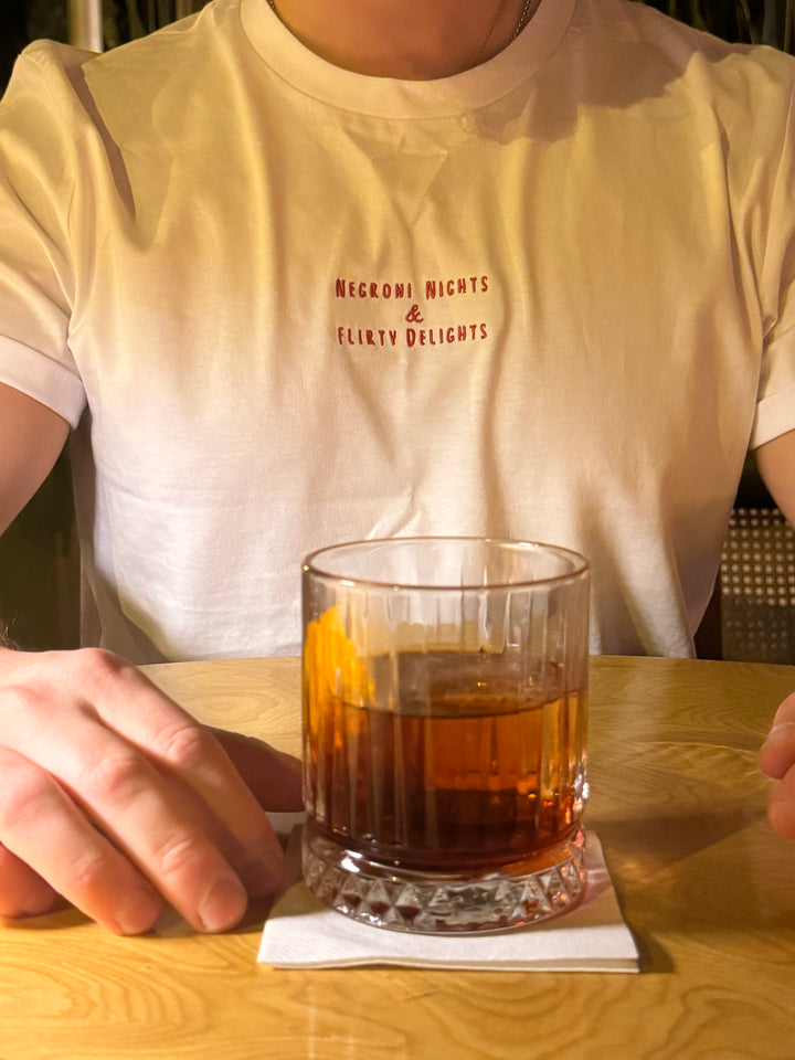 A person wearing a Negroni Nights & Flirty Delights Organic T-shirt, sitting with a negroni cocktail on a table.