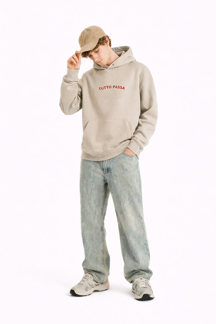 Person wearing a beige hoodie with text, light blue jeans, and a cap on a white background