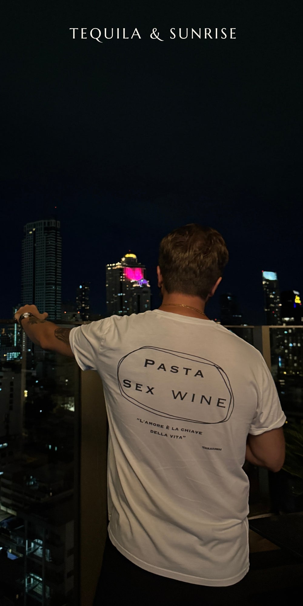 Man wearing Pasta & Sex & Wine Organic T-shirt against a city skyline at night.