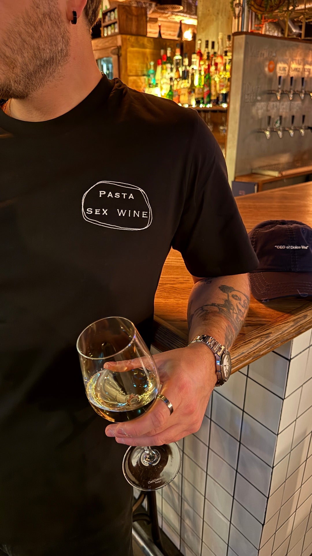Man holding a glass of wine while wearing Pasta & Sex & Wine Organic T-shirt in a bar setting.