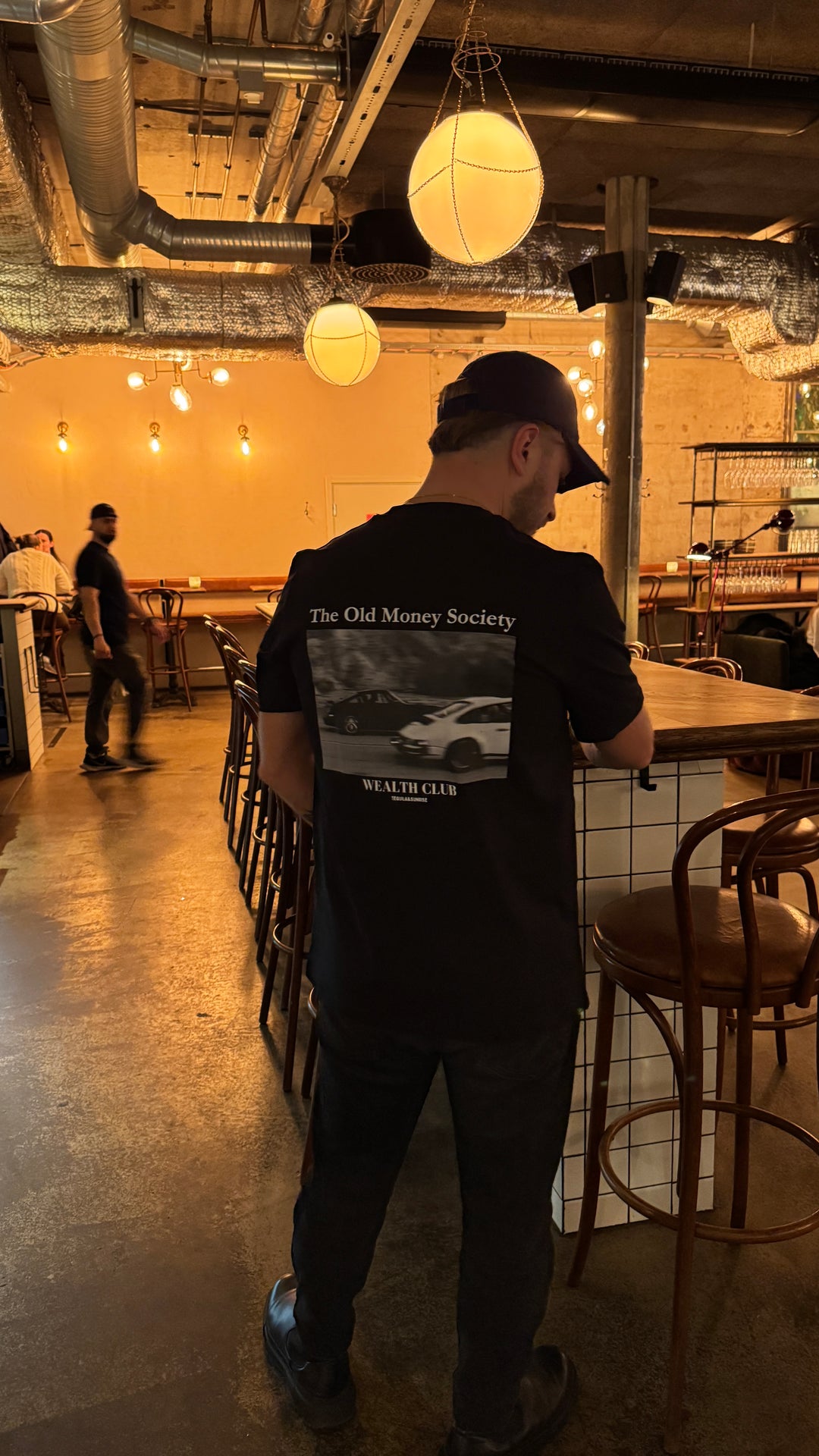 Person wearing an Old Money Society Organic T-shirt in a stylish restaurant setting.