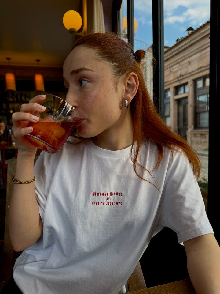 Model enjoying a drink while wearing the Negroni Nights & Flirty Delights Organic T-shirt in a cozy café setting.