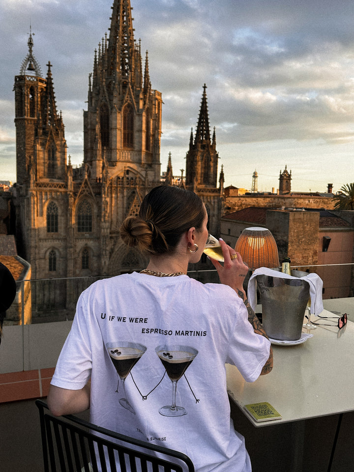 A person wearing the If We Were Espresso Martinis Organic T-shirt, enjoying a cocktail with a cityscape view.