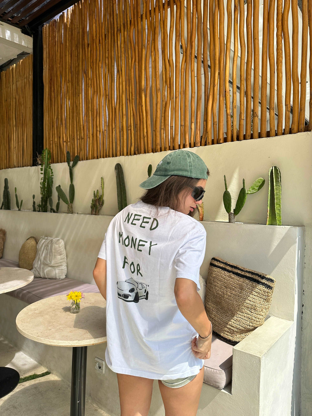 A person wearing a Tequila & Sunrise "NEED MONEY FOR PORSCHE" organic t-shirt stands near a wall adorned with cacti and a woven basket.