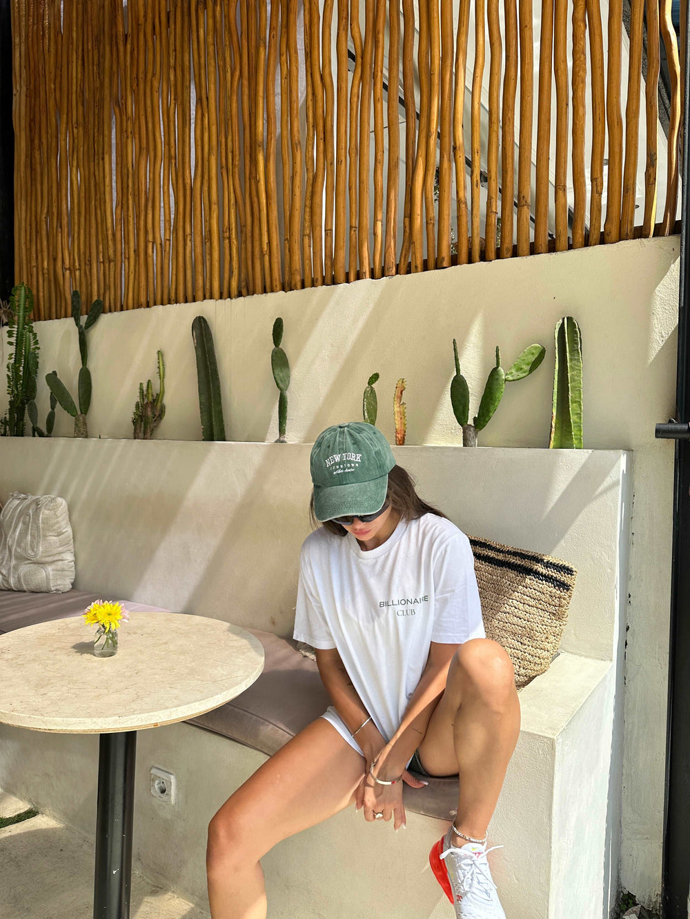 A person in an eco-friendly, white Billionaire Club Minimalist Organic T-shirt by Tequila & Sunrise and a green cap sits on a bench near a table adorned with a yellow flower in a vase. Cacti rest on a ledge, while wooden sticks craft an overhead screen.