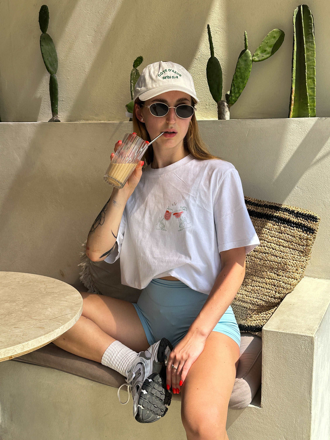 A person in sunglasses and a cap relaxes on a cushioned bench, sipping from a straw. They sport "The Aperol Spritz Club Organic T-shirt" by Tequila & Sunrise, perfectly blending style with sustainability. Cacti stand tall in the background, complementing the eco-friendly vibe.