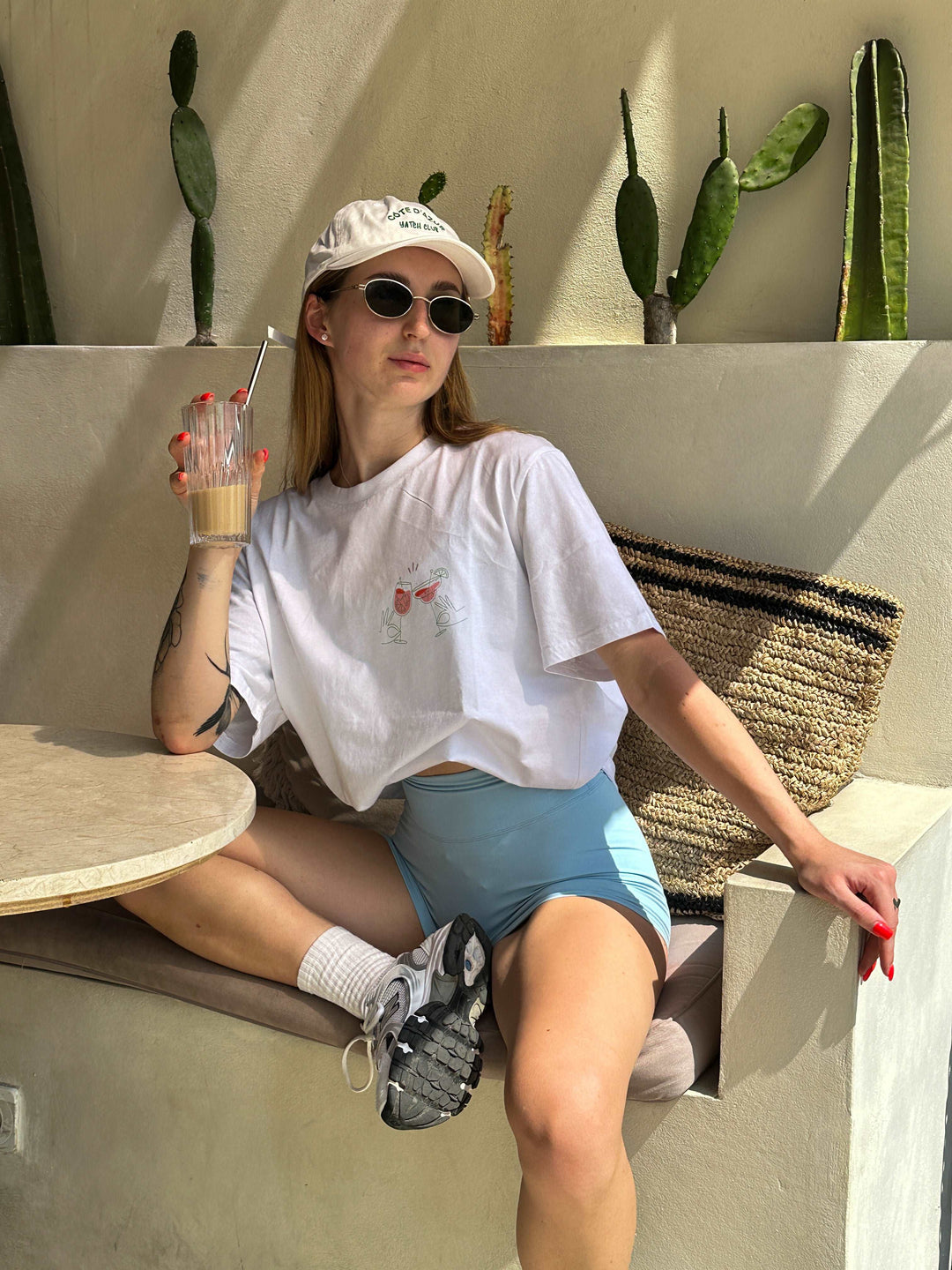 A person wearing The Aperol Spritz Club Organic T-shirt by Tequila & Sunrise, along with blue shorts and sunglasses, sits on a bench surrounded by cactus plants, sipping a drink through a straw.