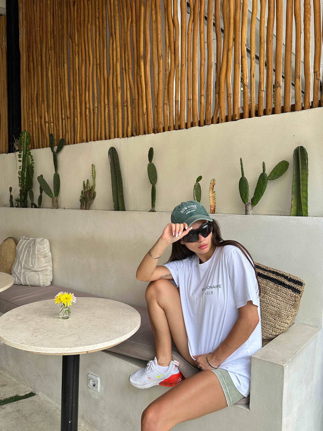 A person relaxes on a bench wearing a casual NEED MONEY FOR PORSCHE ORGANIC T-SHIRT from Tequila & Sunrise, pairing it with a cap and sunglasses. They sit near a table adorned with a small vase of yellow flowers. Behind them, cacti align the wall, enhanced by the bamboo railing above.