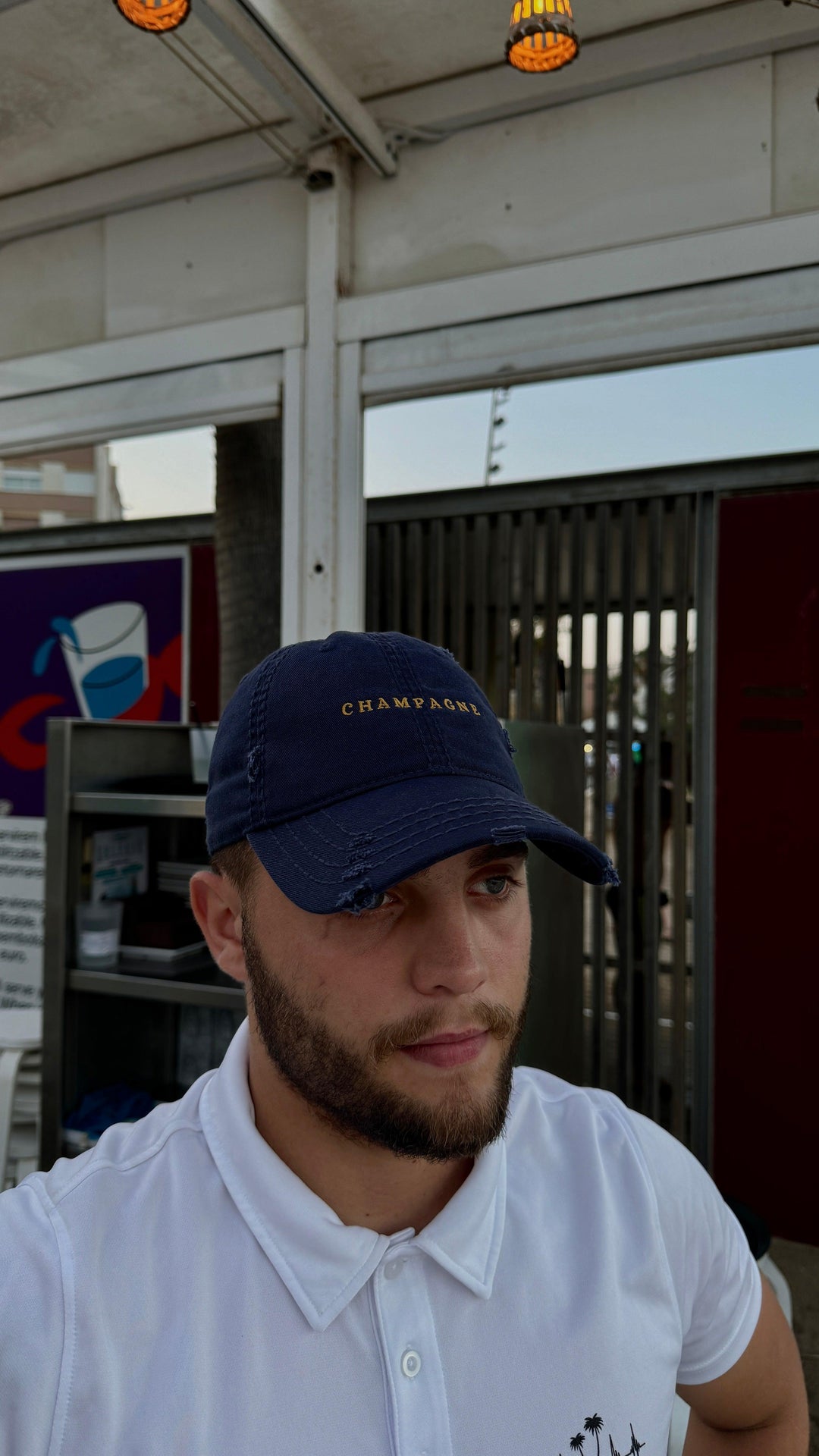 A bearded man, dressed in a sophisticated outfit, wears "The Champagne Hat" by Tequila & Sunrise—a navy dad hat featuring "CHAMPAGNE"—along with a crisp white shirt as he stands in front of a metal fence and signage.