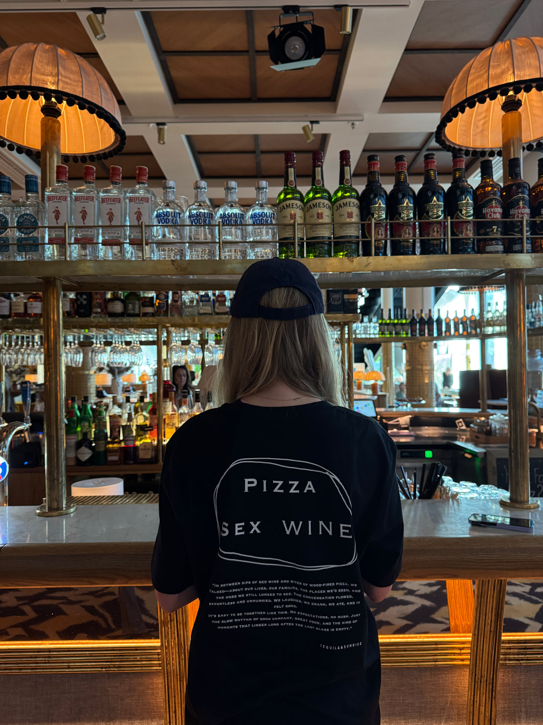 Person wearing a Pizza & Sex & Wine Organic T-shirt at a stylish bar with bottles displayed.