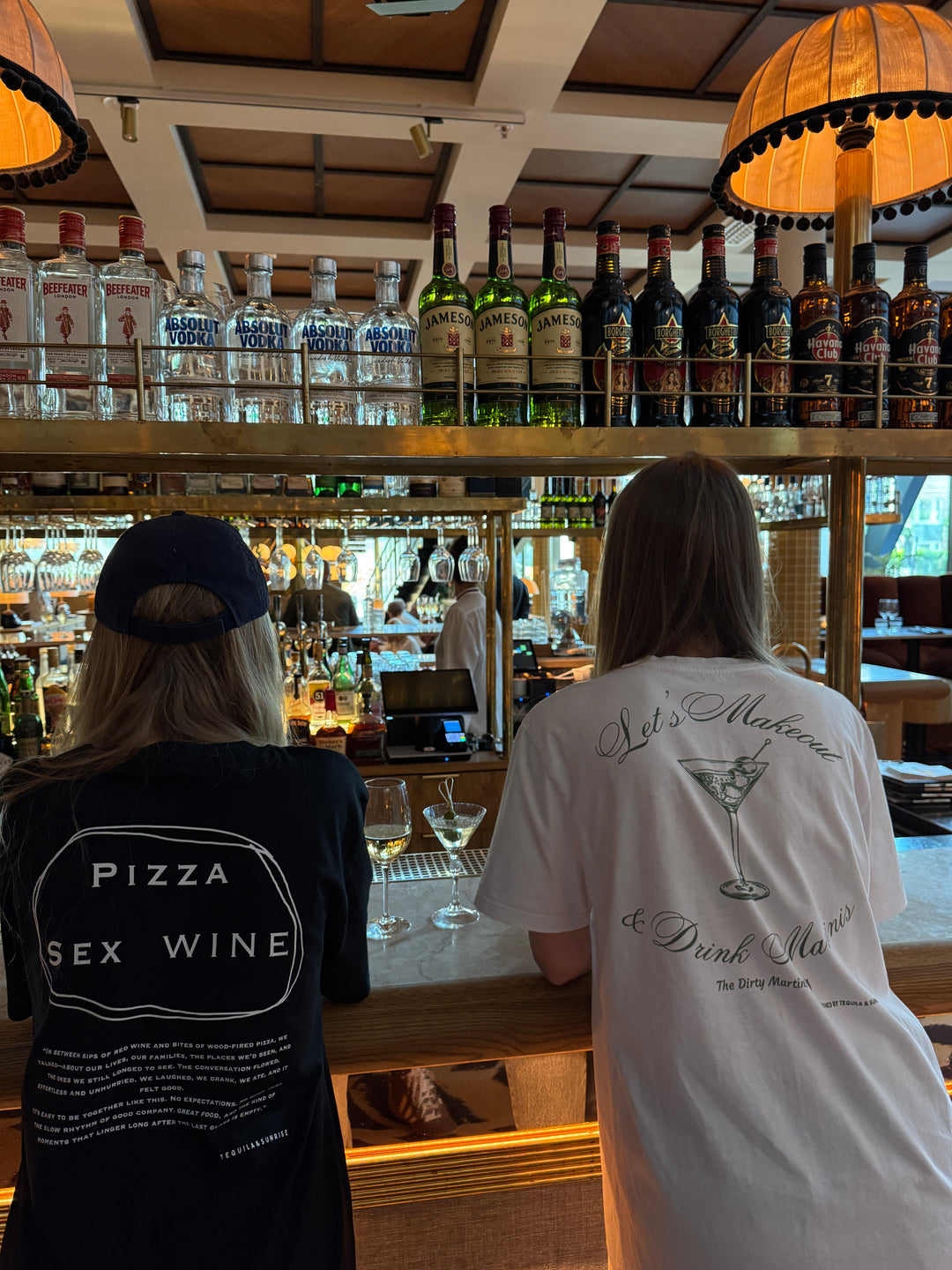 Two people at a bar wearing Pizza & Sex & Wine Organic T-shirt and a white t-shirt with a cocktail design.