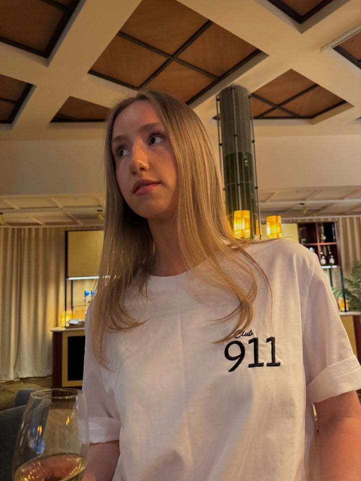 Model wearing a Porsche Club 911 Organic T-shirt in a stylish indoor setting, holding a glass of wine.