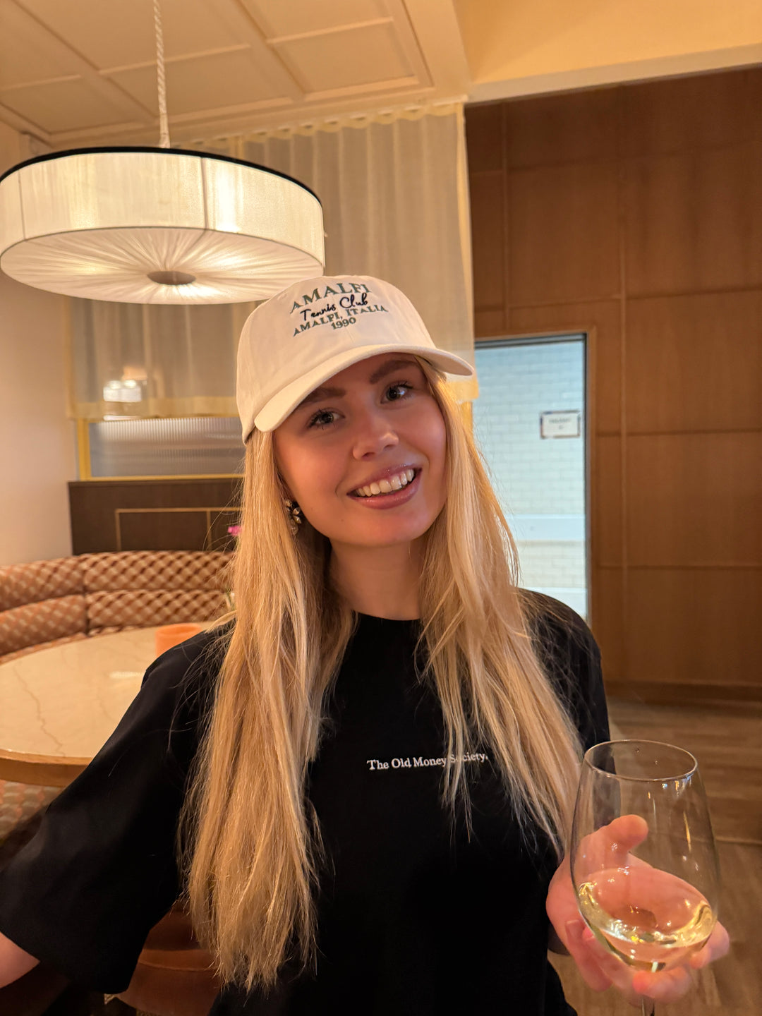 Model wearing The Old Money Society Organic T-shirt and a cap, holding a glass of wine, smiling.