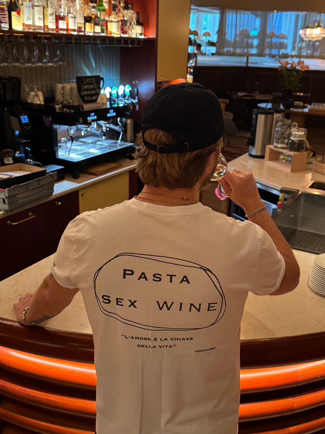 Person wearing a Pasta & Sex & Wine Organic T-shirt at a bar, enjoying a drink, highlighting Italian culture.