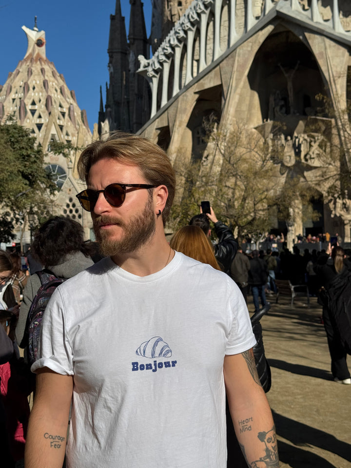 Man wearing sunglasses and a white t-shirt with 'Bonjour' design in front of a large architectural structure.