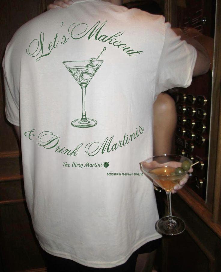 A member of the Naughty Martinis Club is seen holding a cocktail glass while wearing a Tequila & Sunrise 'Let's Makeout & Drink Martinis' Organic T-shirt, crafted from eco-friendly organic cotton and featuring a playful martini design.