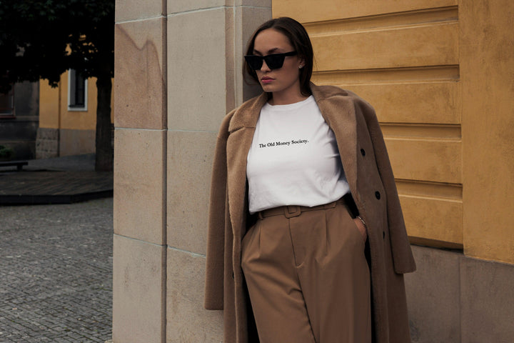 A woman in sunglasses leans against a building, wearing The Old Money Society Organic T-shirt by Tequila & Sunrise, complemented by a tan coat and trousers.