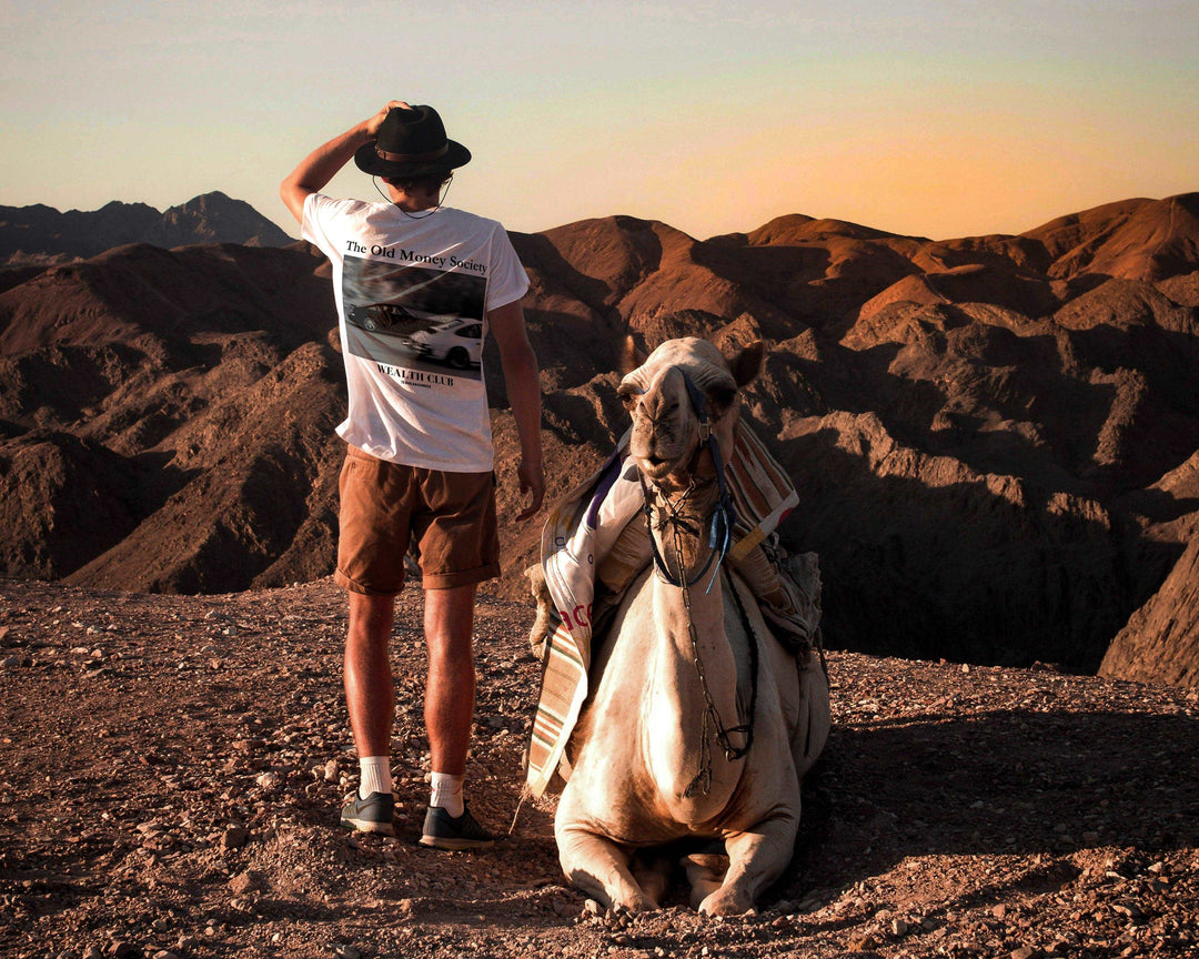 Person wearing Old Money Society Organic T-shirt standing beside a camel in a scenic desert landscape.