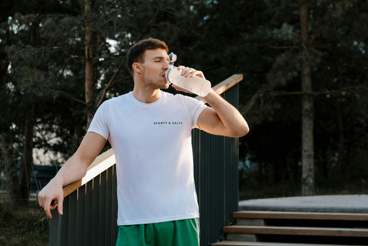 A fit man wearing a Sporty & Salty Organic T-shirt, drinking water outdoors, showcasing active lifestyle.