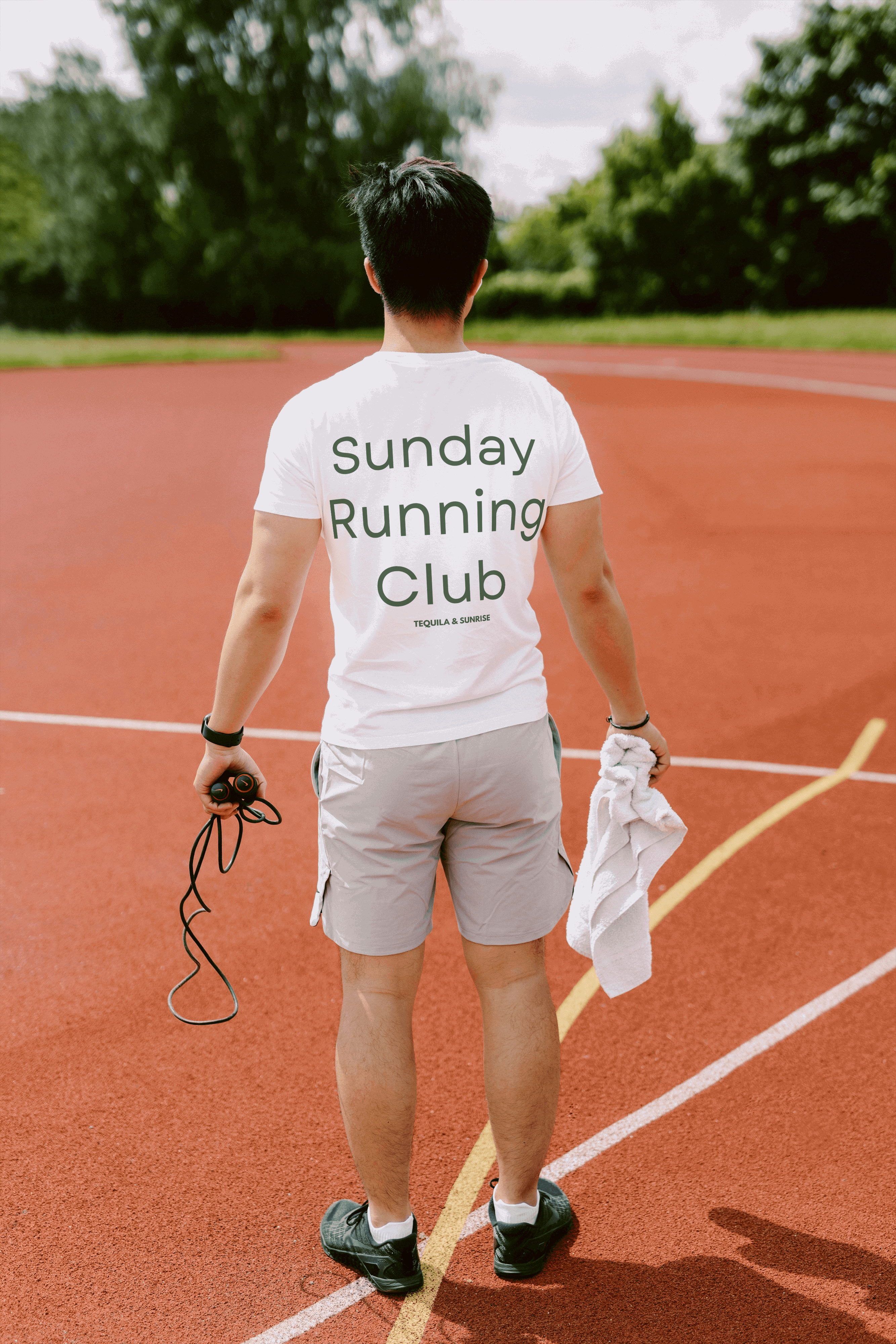 A person stands on a track, holding a jump rope and towel. They wear an organic cotton t-shirt from Tequila & Sunrise’s Sunday Running Club collection, blending eco-friendly style with their active spirit.