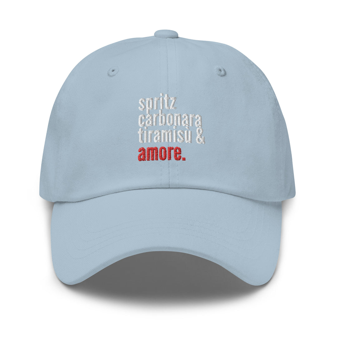 The Love from Italy Hat by Tequila & Sunrise is a light blue dad hat made from chino cotton twill, featuring an adjustable strap and embroidered text: spritz carbonara tiramisu & amore, with amore highlighted in red.