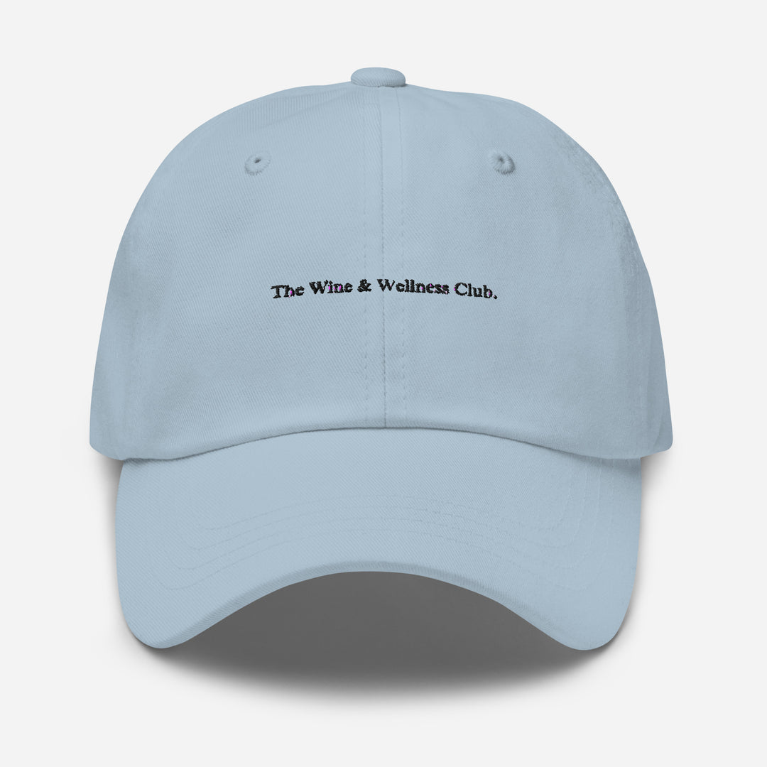 The Wine & Wellness Club by Tequila & Sunrise is a light blue hat crafted from chino cotton twill, showcasing black text that reads "The Wine & Wellness Club." It features an adjustable strap for a perfect fit.