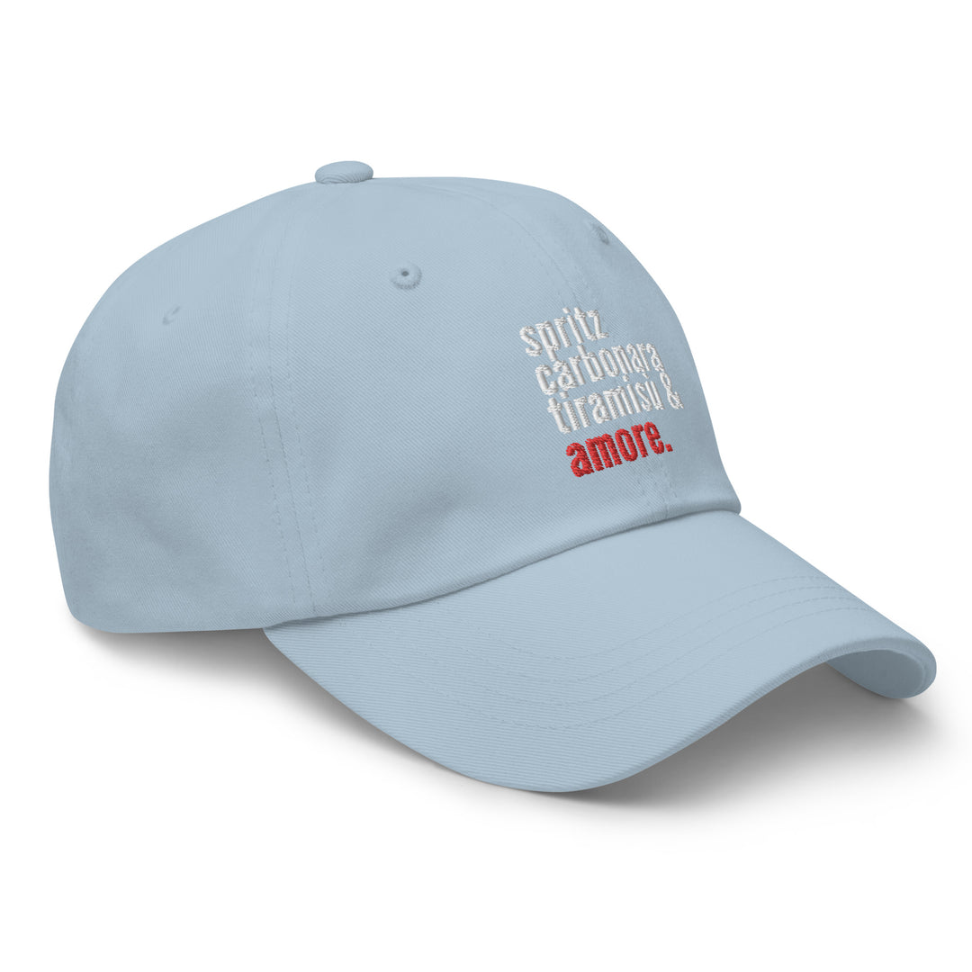 The Love from Italy Hat by Tequila & Sunrise is a light blue dad hat made from chino cotton twill. It showcases the words "spirit," "carbonara," "tiramisu," and "amore" embroidered in white and red on the front, and it also features an adjustable strap for a perfect fit.