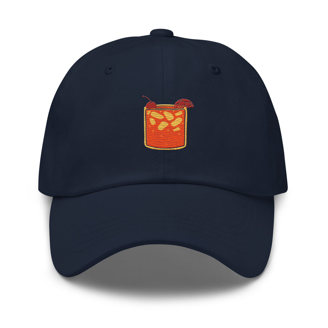 The Tequila & Sunrise Glass Hat by Tequila & Sunrise is a dark blue Dad hat with a low-profile design, showcasing an embroidered orange drink complete with ice cubes and a straw on the front. It features an adjustable strap for a perfect fit.