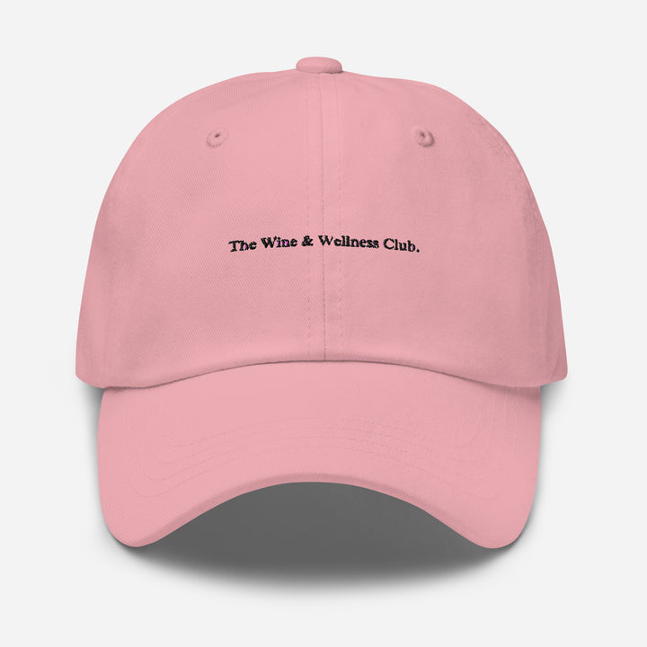 The Wine & Wellness Club by Tequila & Sunrise is a pink hat crafted from chino cotton twill, showcasing black text that reads "The Wine & Wellness Club." It features an adjustable strap for a perfect fit.