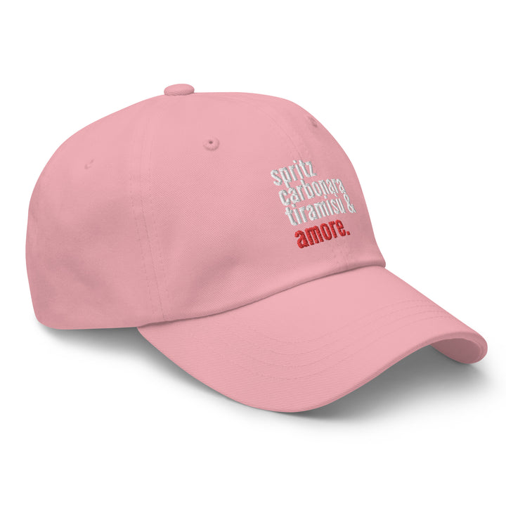 The Love from Italy Hat by Tequila & Sunrise is a pink dad hat made from chino cotton twill, adorned with embroidered text in white and red: spritz, carbonara, tiramisu & amore. It features an adjustable strap for the perfect fit.