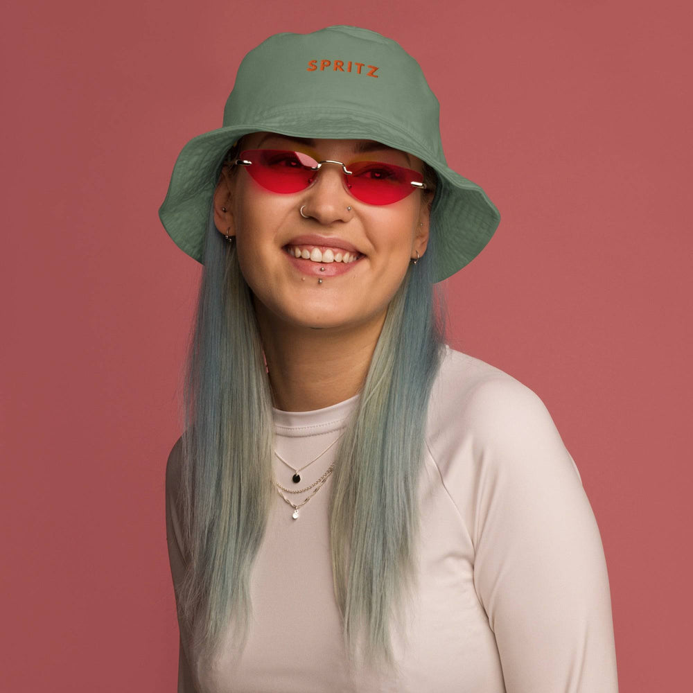 A person with blue hair wearing Tequila & Sunrise's The Spritz Organic Bucket Hat in green, along with red oval glasses and a 100% cotton white shirt, smiles against a pink background.