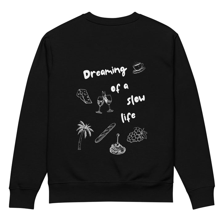 The La Vita Lenta Eco Sweatshirt by Tequila & Sunrise is a black, durable piece crafted from organic cotton. It features white illustrations of a coffee cup, cheese wedge, wine glasses, palm tree, baguette, stack of pancakes, and grapes. The text whimsically reads "Dreaming of a slow life.