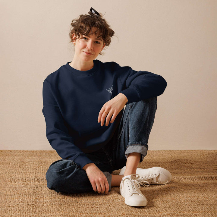 A person with curly hair is sitting cross-legged on a woven rug, dressed in the Let's Makeout & Drink Martinis Eco Sweatshirt by Tequila & Sunrise, paired with blue jeans and white sneakers. They're part of the sustainable fashion trend championed by the Naughty Martinis Club.