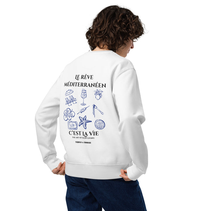 A person with curly hair is wearing "The Mediterranean Dream Eco Sweatshirt" by Tequila & Sunrise, crafted from organic cotton. Adorned with illustrations and French text featuring elements like a seashell, sunglasses, and a glass, they pose against a plain background, perfectly capturing the spirit of slow living.