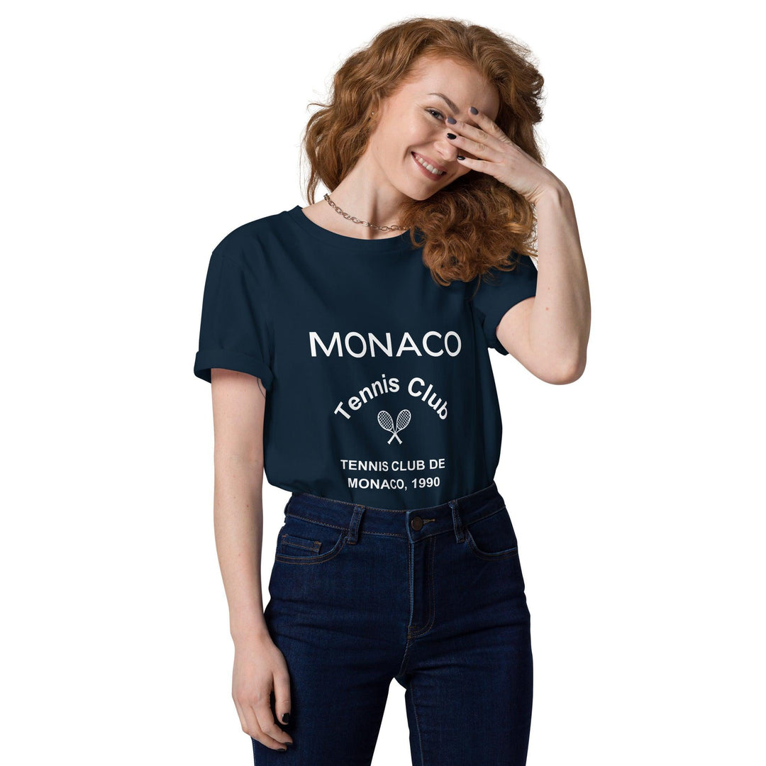 A person with long red hair, wearing an eco-friendly navy blue Tequila & Sunrise Monaco Tennis Club Organic T-shirt and jeans, is smiling while partially covering their face with one hand.