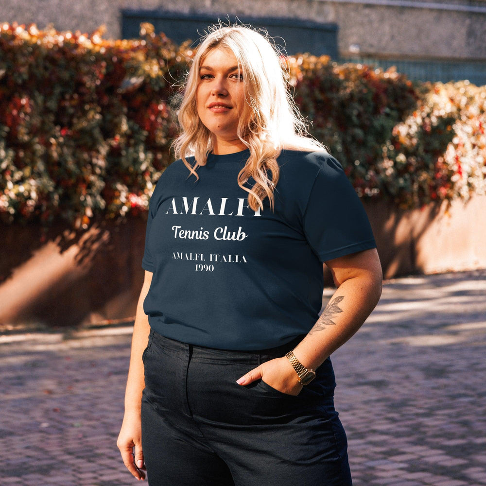 A person stands outdoors wearing a Tequila & Sunrise Amalfi Tennis Club Summer Italia 1980 Organic T-shirt, surrounded by vibrant autumnal foliage.