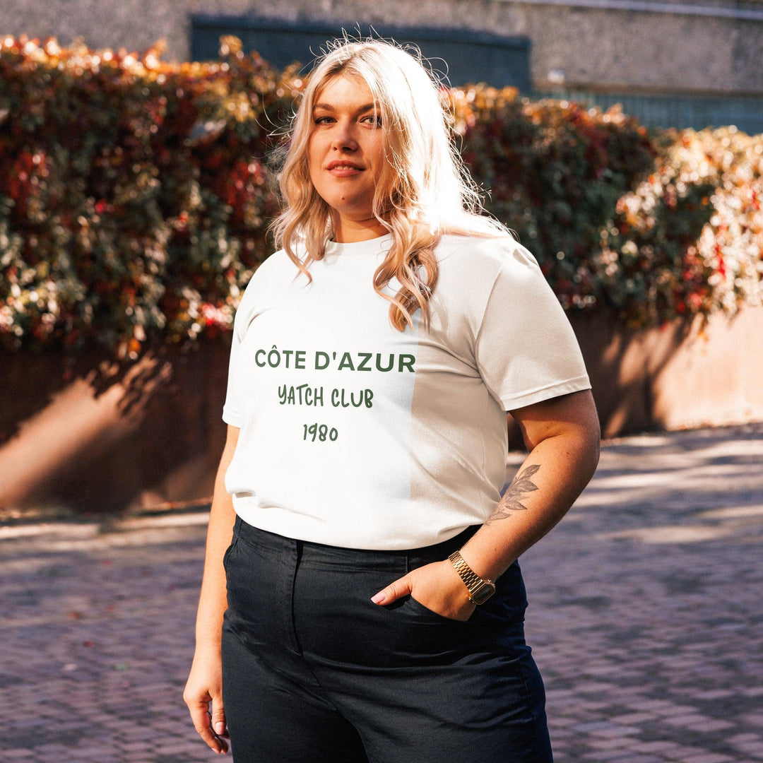 In the sunlight, a person with long hair stands outdoors wearing an eco-friendly Côte d'Azur Yacht Club Organic T-shirt by Tequila & Sunrise and dark pants, capturing the relaxed elegance of the French Riviera amidst a backdrop of lush foliage.