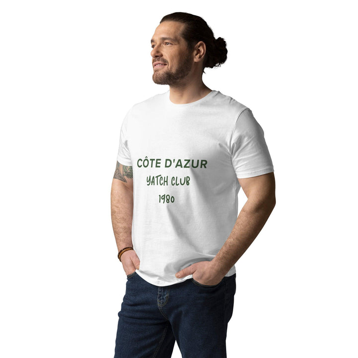 A man with a ponytail and tattoos is wearing a Tequila & Sunrise Côte d'Azur Yacht Club Organic T-shirt, effortlessly showcasing the laid-back elegance of the French Riviera while standing with one hand in his pocket.