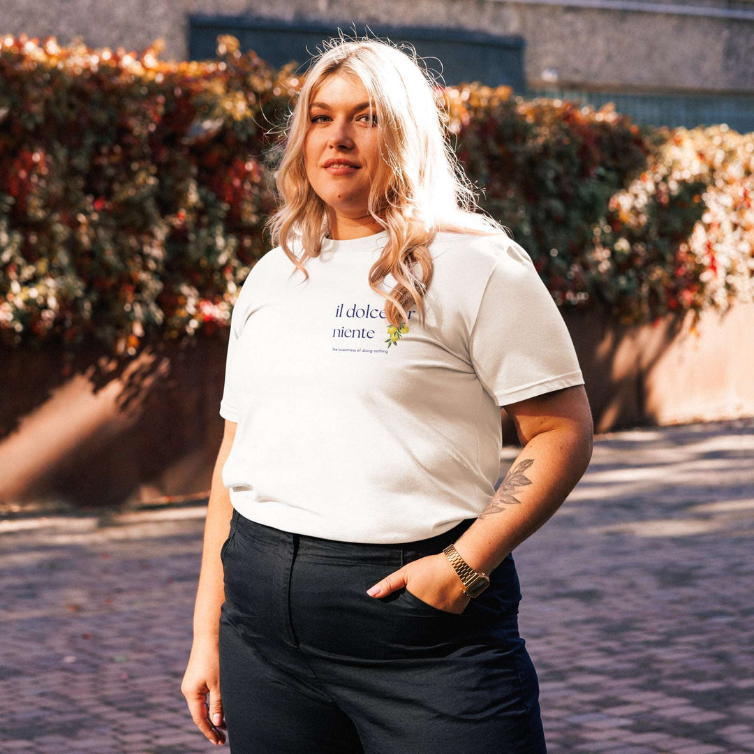 A person with long blonde hair stands outdoors, embracing the Mediterranean lifestyle in an eco-friendly The Il Dolce Far Niente Organic T-shirt by Tequila & Sunrise and black pants. The vibrant hues of colorful leaves create a picturesque backdrop.